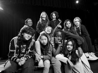 Black-and-white photo of a group of young people seated and standing together on a theatre stage, some holding saxophones, facing the camera under stage lighting.