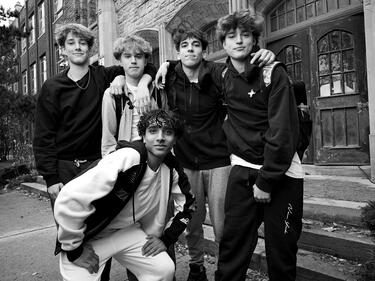 five male students standing gathered together outside of their school. 